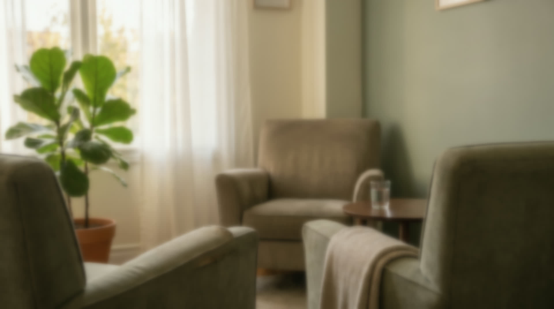 A quiet room with two soft chairs, a blanket over one arm, a glass of water on a low table, and morning light through a curtain.